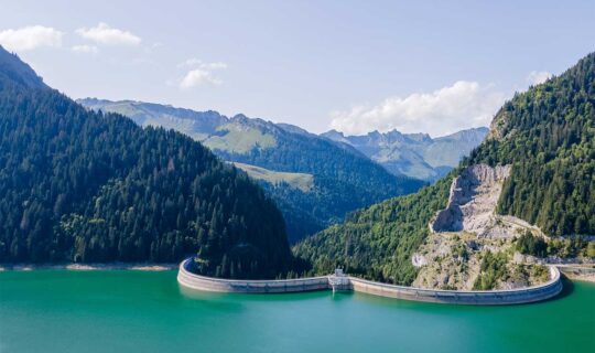 Strommangellage: einzig kurzfristige Entwarnung. Bild zeigt einen Staudamm in den Schweizer Voralpen mit zur Zeit hohem Wasserstand. Das strukturelle Problem der Schweiz bleibt für die kommenden Jahre bestehen und kann angebotsseitig nicht kurzfristig behoben werden. Deshalb sind die Unternehmen gut beraten, sich weiterhin gut und umfassend vorzubereiten.